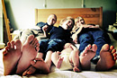 A family lie together in bed laughing with their different sized bare feet towards the camera