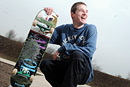 A young man poses with his skateboard with a skate park as the backdrop